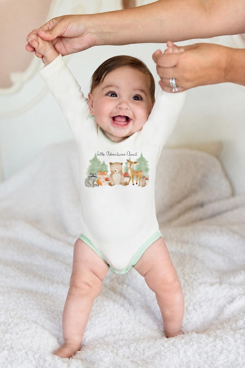 Puede incluir: Un body de beb&eacute; de manga larga en color crema con ribetes verde claro. La parte delantera presenta un gr&aacute;fico de acuarela de un mapache, zorro, oso, ciervo y conejo en un bosque con el texto Little Adventures Await impreso sobre ellos.