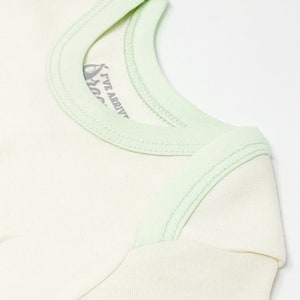 Puede incluir: Una camiseta de color crema con ribete verde menta claro a lo largo del cuello y las costuras de los hombros. La etiqueta interior del cuello presenta un gr&aacute;fico de un aguacate y el texto I'VE ARRIVED en gris oscuro, visible por el cuello.