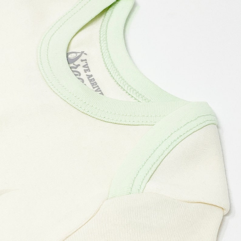 Puede incluir: Una camiseta de color crema con ribete verde menta claro a lo largo del cuello y las costuras de los hombros. La etiqueta interior del cuello presenta un gr&aacute;fico de un aguacate y el texto I'VE ARRIVED en gris oscuro, visible por el cuello.