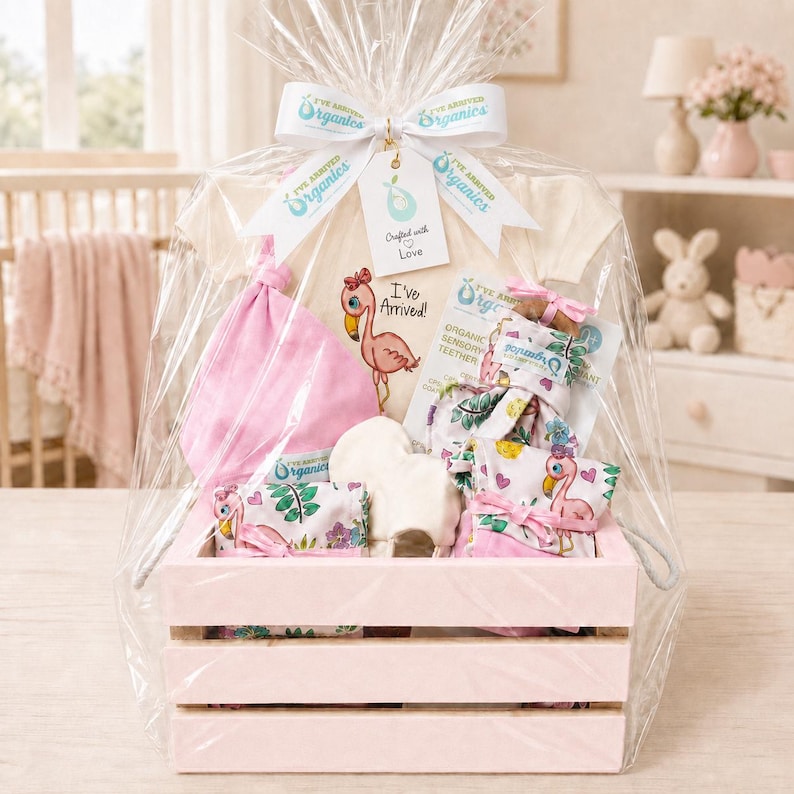 May include: A baby gift set in a light pink wooden crate wrapped in clear cellophane. It includes a cream-colored onesie with a flamingo graphic and the text I've Arrived!, a pink hat, a white cloth, and a sensory teether with a flamingo print.
