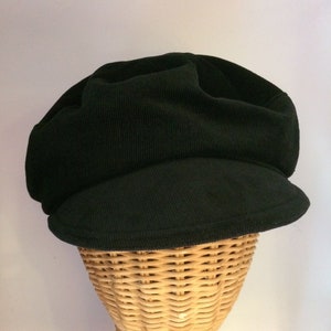 Puede incluir: Una gorra de estilo newsboy de pana negra con una corona ligeramente redondeada y una visera que se curva hacia abajo en los lados. La gorra se muestra en una cabeza de maniquí de mimbre.