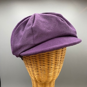 May include: A purple newsboy cap with a rounded crown and a short, stiff brim. The cap is made of a soft, textured fabric and is displayed on a wicker head form. The cap's color is a deep, rich shade of purple.
