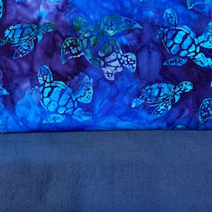 May include: A fabric design featuring sea turtles in shades of blue and green against a dark blue and purple background. The turtles have detailed patterns on their shells. The lower portion of the image is a solid, dark blue fabric.