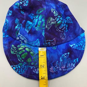 May include: A blue fabric hat with a pattern of sea turtles in shades of blue and green. The hat is approximately 5 inches in height. The hat has a curved brim and is made of a soft fabric.