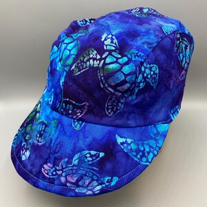 May include: A blue baseball cap with a sea turtle pattern. The cap features a vibrant design of turquoise and white sea turtles against a deep blue background. The brim and crown are fully covered in the turtle print.