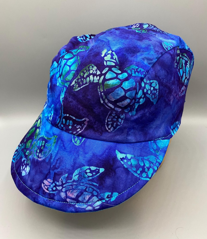 May include: A blue baseball cap with a sea turtle pattern. The cap features a vibrant design of turquoise and white sea turtles against a deep blue background. The brim and crown are fully covered in the turtle print.