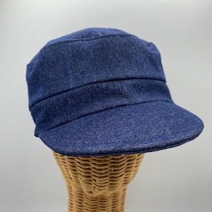 May include: A dark blue denim cap with a short brim. The cap has a structured design with a horizontal seam across the front. The hat is displayed on a wicker form.