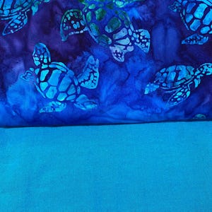 May include: A fabric with a blue and teal color scheme. The top portion features a pattern of sea turtles in shades of blue and green against a dark blue background. The bottom portion is a solid teal color.