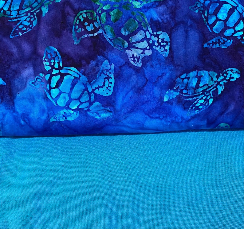May include: A fabric with a blue and teal color scheme. The top portion features a pattern of sea turtles in shades of blue and green against a dark blue background. The bottom portion is a solid teal color.
