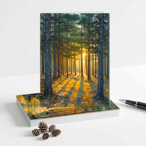 May include: A greeting card featuring a forest scene with tall trees and sunlight streaming through the branches. The artwork depicts a vibrant landscape with a warm, golden glow, creating a serene and natural ambiance. The card is resting on a stack of white paper.