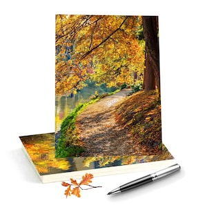 May include: A rectangular greeting card featuring a vibrant autumn forest path with golden yellow and orange leaves, displayed alongside a silver metal ballpoint pen and a small, detached brown oak leaf resting on a plain white surface.