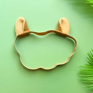 May include: A tan elastic headband featuring two small, upright, rounded felt ears attached to the top. The headband is constructed from a thin, flexible, light brown material that forms a circular shape, designed to sit comfortably around the head.