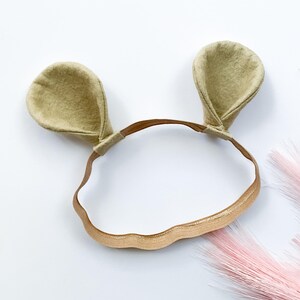 May include: A light tan headband with two matching felt ears. The headband is a thin, gold elastic band. The ears are a rounded shape and are attached to the headband. The background is white with pink decorative elements.