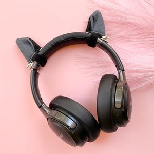 May include: Black over-ear headphones with gray cat ear attachments. The headphones are on a pink background with a fluffy pink texture in the background. The headphones have a sleek, modern design.