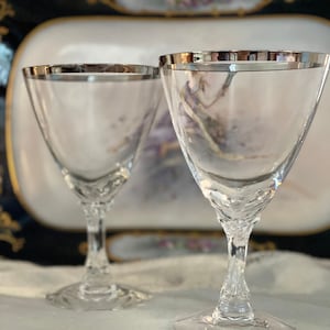Peut inclure: Deux verres à vin en verre transparent avec des bords argentés sur une nappe en dentelle blanche.