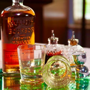 May include: A still life arrangement featuring a bottle of Bulleit Bourbon Frontier Whiskey, clear shot glasses, and decorative bottles. The items are arranged on a green mirrored tray, creating a classic bar setup.