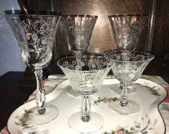 May include: Set of five clear glass wine glasses with etched floral designs. The glasses are arranged on a white tray with a floral pattern.