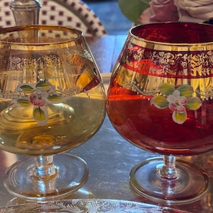 May include: Two vintage glass decanters with gold trim and painted floral designs. One decanter is amber glass with a white floral design, and the other is red glass with a white and green floral design.