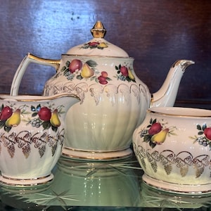May include: A vintage tea set comprising a teapot, creamer and sugar bowl. The set is decorated with a floral pattern featuring pears, apples and grapes. The background is a pale green colour with gold accents.