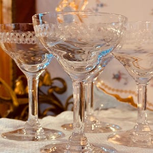 May include: Four clear, stemmed glasses with etched detailing around the rims. The glasses have a classic design with a rounded bowl and a slender stem. The background is softly blurred, suggesting a dining setting.