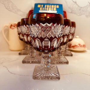 May include: A set of deep red cut-to-clear crystal wine glasses featuring a diamond-patterned upper bowl, a smooth lower bowl, a clear glass stem with a spherical node, and a square base with a sunburst design radiating from the centre.
