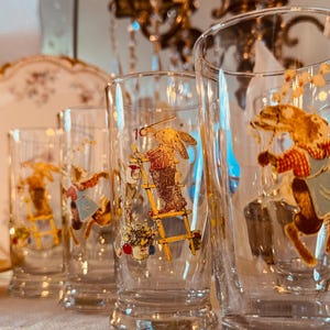 May include: Set of clear drinking glasses with a whimsical design featuring a brown horse in a red shirt and blue apron. The glasses are decorated with gold accents and are part of a vintage glassware collection.
