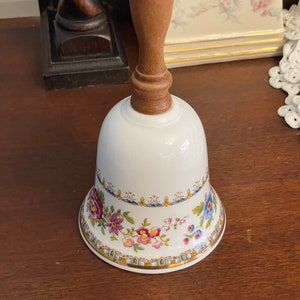 May include: A white porcelain bell with a floral design and a wooden handle. The bell has a gold trim and blue and pink flowers.