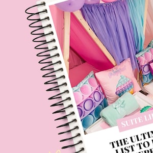 May include: A pink notebook with a black spiral binding is in the foreground. The background features a play tent with pink, purple, and blue decor, including pillows and a blanket. The pillows have a pop-it design.