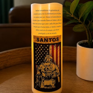 May include: A cylindrical candle with a patriotic design. The candle features the American flag, a soldier illustration, and the name "SANTOS". The candle has text in Spanish. The candle is on a wooden surface.