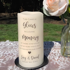 May include: A cream-colored pillar candle with the words "This Candle Glows In Loving Memory" printed in elegant script. The candle includes the names "Lucy & David" and the date August 24, 2023. A black metal base supports the candle.