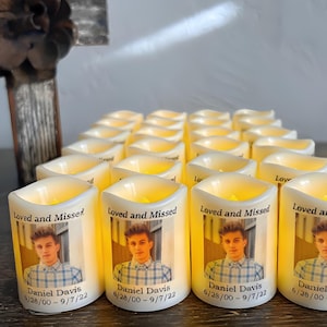 Op de afbeelding: Een verzameling witte LED-stompkaarsen met een afgedrukte foto van een jong persoon in een geruit overhemd. Elke kaars toont de tekst Loved and Missed, de naam Daniel Davis en de data 6/28/00 - 9/7/22 in zwarte letters.