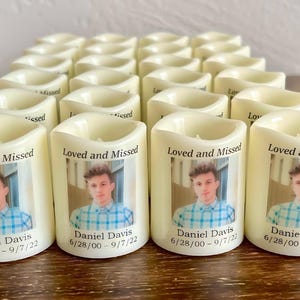 Op de afbeelding: Een verzameling gebroken witte, cilindervormige LED-herdenkingskaarsen met een afgedrukte foto van een jong persoon in een blauw geruit overhemd. Elke kaars toont de tekst Loved and Missed, de naam Daniel Davis en de data 6/28/00 - 9/7/22.