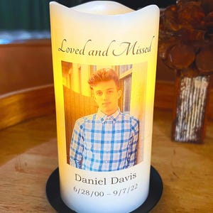 May include: A white LED candle with a photo of a young man in a blue plaid shirt. The candle has the text "Loved and Missed" and "Daniel Davis 6/28/00 - 9/7/22". The candle sits on a black base.