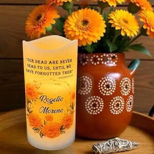 K&ouml;nnte beinhalten: Eine leuchtende Kerze mit dem Zitat "OUR DEAD ARE NEVER DEAD TO US, UNTIL WE HAVE FORGOTTEN THEM" und dem Namen "Rogelio Moreno". Die Kerze ist mit orangefarbenen Blumen verziert. Eine Vase mit orangefarbenen Blumen und ein dekorativer Topf befinden sich im Hintergrund.