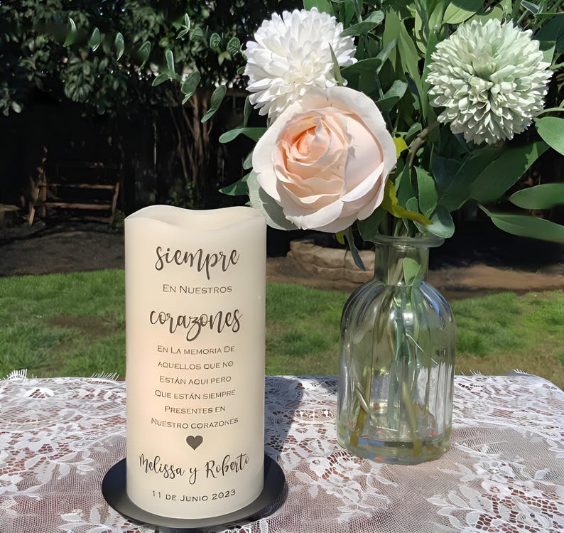 May include: A cream pillar candle with the Spanish text "Siempre en nuestros corazones" sits on a black base. A floral arrangement with a peach rose and white flowers is in a glass vase. The candle is on a lace tablecloth.