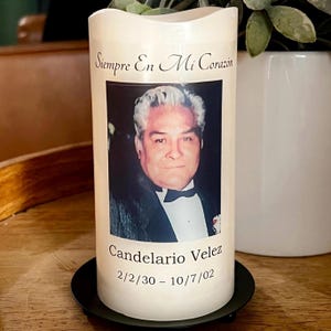 May include: Cream-colored pillar candle with a photograph of a man in a tuxedo. The candle has the text "Siempre En Mi Corazón" above the photo, and "Candelario Velez" with dates 2/2/30 - 10/7/02 below the photo. The candle sits on a black metal base.