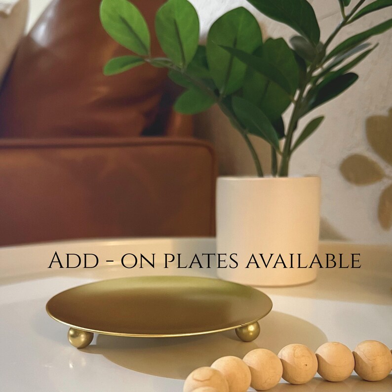 May include: A gold-coloured, round plate with three small, spherical feet. The plate is on a white surface, with the text "ADD - ON PLATES AVAILABLE" in the image. A small, white cylindrical planter with a green plant is in the background.