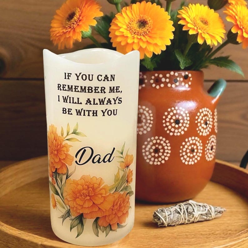 K&ouml;nnte beinhalten: Eine wei&szlig;e Kerze mit dem Text "IF YOU CAN REMEMBER ME, I WILL ALWAYS BE WITH YOU" und dem Wort "Dad". Die Kerze ist mit orangefarbenen floralen Designs verziert. Eine Vase mit orangefarbenen Blumen und ein dekorativer Topf befinden sich im Hintergrund.
