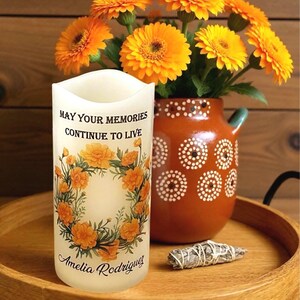 K&ouml;nnte beinhalten: Eine wei&szlig;e Kerze mit dem Text "MAY YOUR MEMORIES CONTINUE TO LIVE" und "Amelia Rodriguez", umgeben von einem Blumenkranz. Im Hintergrund eine Vase mit orangefarbenen Blumen und eine braune Vase mit wei&szlig;en Punkten. Ein Salbeib&uuml;ndel liegt auf einem Holztablett.
