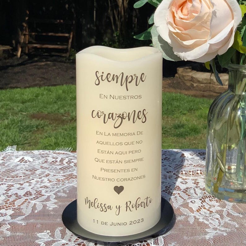 Peut inclure: Une bougie pilier cr&egrave;me avec l'inscription en espagnol "Siempre En Nuestros Corazones". La bougie, pos&eacute;e sur une base noire, pr&eacute;sente un symbole de c&oelig;ur, les noms "Melissa y Roberto" et la date "11 de Junio 2023".