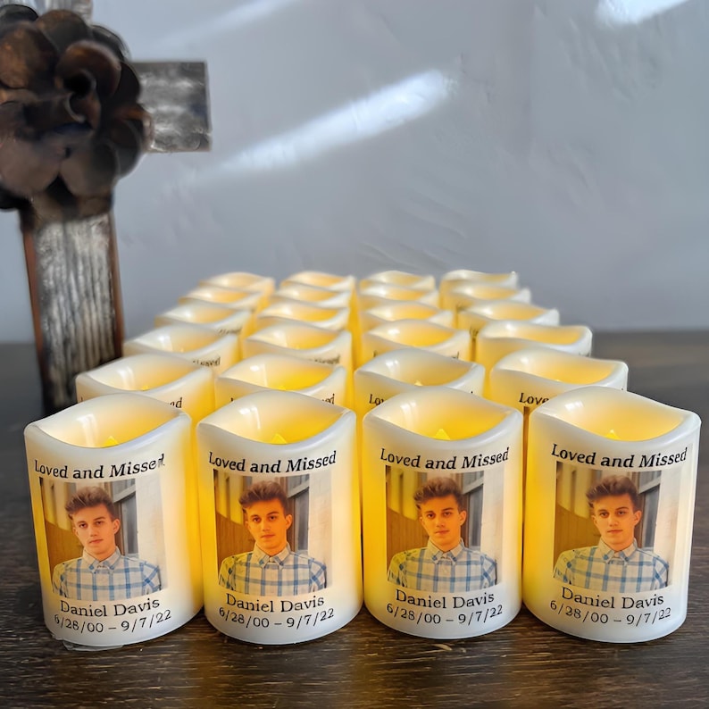 Op de afbeelding: Een verzameling witte LED-stompkaarsen met een afgedrukte foto van een jong persoon in een geruit overhemd. Elke kaars toont de tekst Loved and Missed, de naam Daniel Davis en de data 6/28/00 - 9/7/22 in zwarte letters.