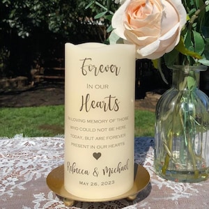 Peut inclure: Une bougie pilier cr&egrave;me portant l'inscription "Forever In Our Hearts" dans une &eacute;criture &eacute;l&eacute;gante. La bougie est personnalis&eacute;e avec les noms "Rebecca & Michael" et la date "26 mai 2023". Elle repose sur une plaque m&eacute;tallique dor&eacute;e, avec un vase de fleurs en arri&egrave;re-plan.