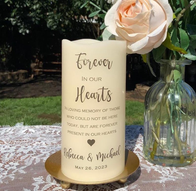 Peut inclure: Une bougie pilier cr&egrave;me portant l'inscription "Forever In Our Hearts" dans une &eacute;criture &eacute;l&eacute;gante. La bougie est personnalis&eacute;e avec les noms "Rebecca & Michael" et la date "26 mai 2023". Elle repose sur une plaque m&eacute;tallique dor&eacute;e, avec un vase de fleurs en arri&egrave;re-plan.