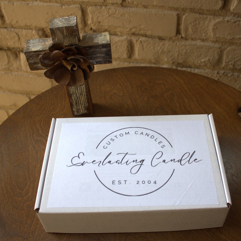 Op de afbeelding: Een witte rechthoekige kartonnen doos met een cirkelvormig logo met de tekst Custom Candles, Everlasting Candle en Est. 2004. Achter de doos staat een rustiek houten kruis versierd met een donkerbruin metalen bloemornament.