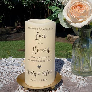Peut inclure: Une bougie pilier cr&egrave;me avec le texte "Because Someone We Love Is In Heaven" et les noms "Mindy & Robert" avec la date du 11 juin 2023. La bougie repose sur une plaque m&eacute;tallique dor&eacute;e. Un vase de fleurs est en arri&egrave;re-plan.