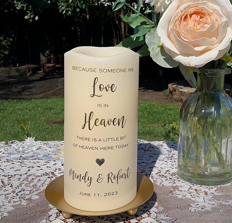 Peut inclure: Une bougie pilier cr&egrave;me avec le texte "Because Someone We Love Is In Heaven" et les noms "Mindy & Robert" avec la date du 11 juin 2023. La bougie repose sur une plaque m&eacute;tallique dor&eacute;e. Un vase de fleurs est en arri&egrave;re-plan.