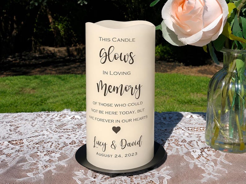 Peut inclure: Une bougie pilier cr&egrave;me avec du texte noir qui dit "This Candle Glows In Loving Memory." La bougie comprend les noms "Lucy & David" et la date "August 24, 2023." La bougie repose sur une base noire, avec un arri&egrave;re-plan flou.