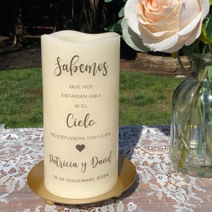Peut inclure: Une bougie pilier cr&egrave;me avec du texte en espagnol, dont le mot "Sabemos", des noms et une date. La bougie est pos&eacute;e sur une plaque m&eacute;tallique dor&eacute;e. Une rose p&ecirc;che et un vase en verre sont en arri&egrave;re-plan.
