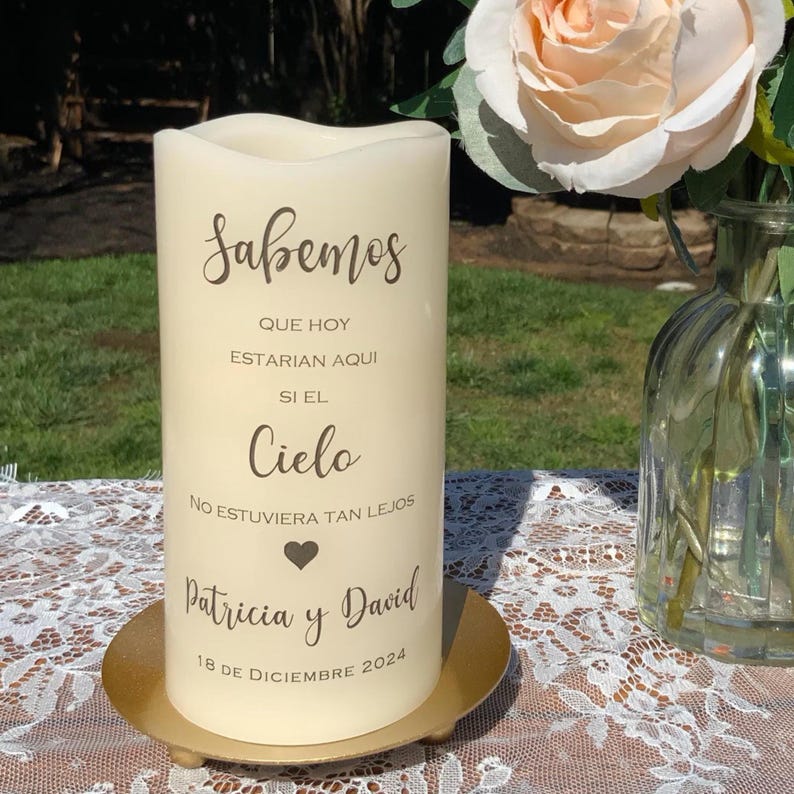 Peut inclure: Une bougie pilier cr&egrave;me avec du texte en espagnol, dont le mot "Sabemos", des noms et une date. La bougie est pos&eacute;e sur une plaque m&eacute;tallique dor&eacute;e. Une rose p&ecirc;che et un vase en verre sont en arri&egrave;re-plan.