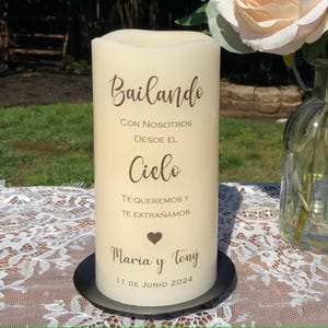 Peut inclure: Une bougie pilier cr&egrave;me avec des inscriptions en marron, dont "Bailando" et "Maria y Tony". La bougie est pos&eacute;e sur un socle noir. La date "11 DE JUNIO 2024" est &eacute;galement visible. Une rose et un vase sont en arri&egrave;re-plan.